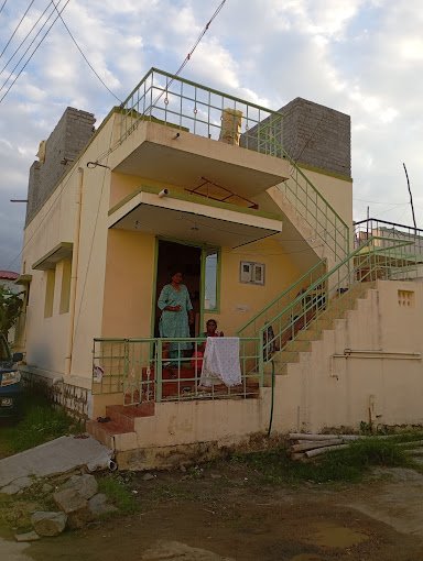 Two-story yellow house with green railings, outdoor staircase, a woman, and child at the entrance, cloudy sky above.