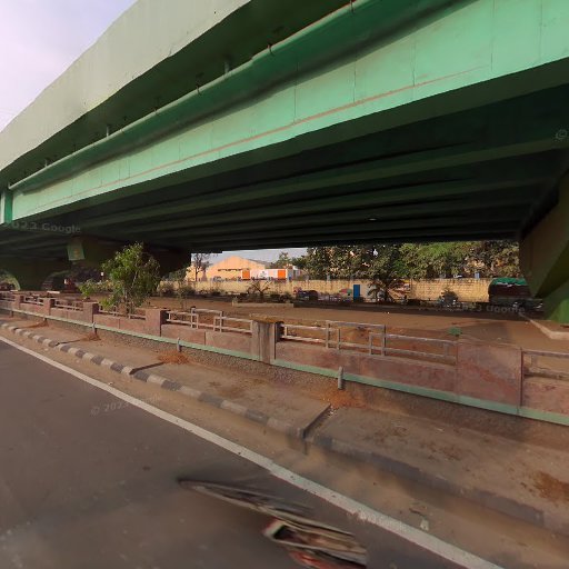 Road under a green overpass with nearby greenery and a few parked vehicles in the background.